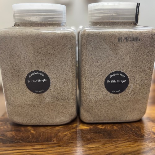 Two square glass jars with white lids, filled with ground brown powder, labeled 'Dr Ellie Wright' on black circular stickers on a wooden surface, side by side.